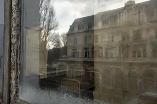 Frosted window of historic thermal building with blurred reflections of hospital beds and archive papers, leafless trees outside, soft winter light.