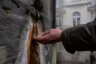 Weathered stone wall in Bains-les-Bains with mineral water trickling down, subtle mineral stains, and an elderly hand gently hovering nearby in soft winter light.